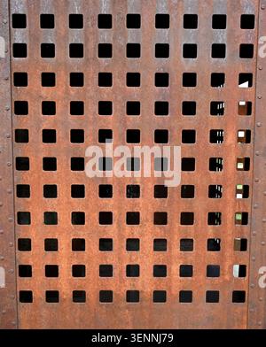 Vertical close up of a rust coated perforated metal panel with square cut out holes and vertical rivets on both sides Stock Photo