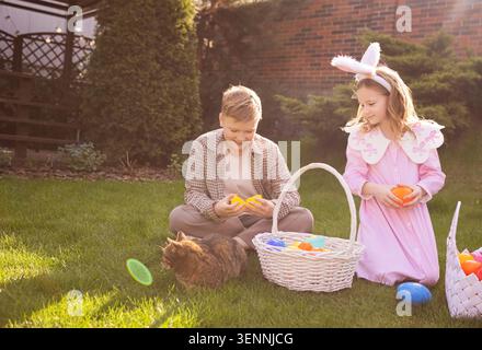 Two children, a boy and a girl, interact with colorful plastic eggs next to a white basket on green grass in a sunny outdoor setting Stock Photo