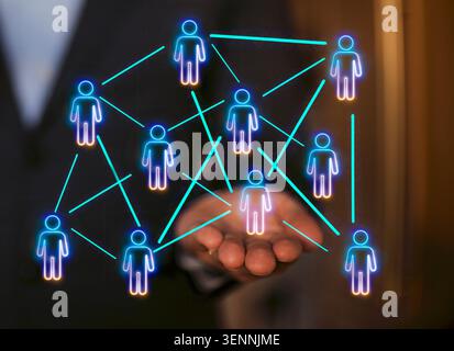 Digital representation of a network with glowing human figures and connecting lines above a businessman's hand in a corporate environment Stock Photo