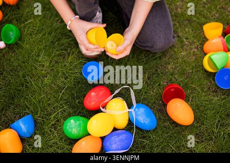 Child's hands are seen holding a yellow plastic egg amidst a variety of colorful plastic eggs scattered on green grass in a festive outdoor setting Stock Photo