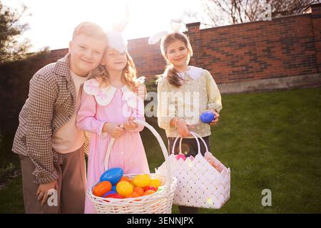 Three kids in festive Easter outfits pose with baskets of colorful eggs in a bright backyard, featuring a brick wall and greenery in the background Stock Photo