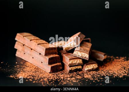 Stacked chocolate wafer bars with cocoa powder on dark background. Sweet dessert snack, milk chocolate pieces with crispy filling, close-up food photo Stock Photo