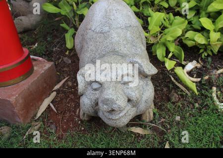 Small stone pig carving resting on grassy ground next to red post at outdoor temple premises. Symbolic animal sculpture. Stock Photo