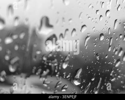 Abstract monochrome close-up of raindrops and water droplets on a glass window pane with a blurred background, creating a moody, melancholy, and peace Stock Photo