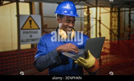 Man in blue hardhat and yellow earmuffs typing on laptop with gloved hands in building; confidence productivity. Stock Photo