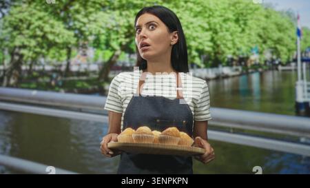 Woman baker holding tray of muffins with both hands on a street by a canal, presenting pastries; curious hospitality. Stock Photo