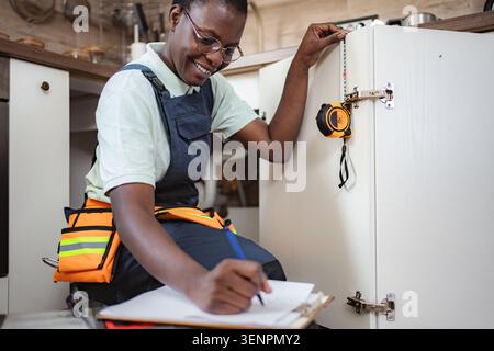 Smiling african american female plumber taking notes and measuring kitchen cabinet Stock Photo