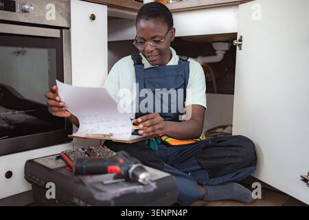 Female plumber checking notes while sitting on kitchen floor Stock Photo