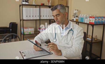 Man doctor with grey hair and stethoscope holds tablet and pen, reviewing clipboard paperwork while seated at desk in medical clinic building; profess Stock Photo