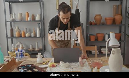 Man leans over laptop with hands on table among clay pieces and pottery tools in studio; concentration craft planning. Stock Photo