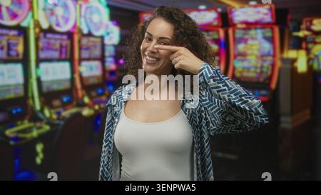 Woman smiling with visible cleavage in a casino building amid colorful slot machines, wearing white tank top and patterned open shirt; happiness luck. Stock Photo