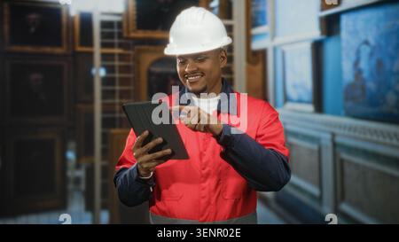 Man in hard hat and red uniform tapping a tablet with finger inside a building, pointing gesture during inspection of gallery paintings; confidence. Stock Photo