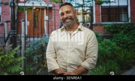Man smiling with hands clasped, wearing linen shirt with rolled sleeves at building entrance outdoors; quiet contentment. Stock Photo