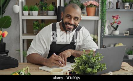 Man florist in apron writes in notebook by laptop at studio desk, pen in hand; calm business planning. Stock Photo