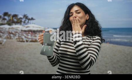 Woman holding moka pot and blowing a kiss toward camera in studio beach set while wearing striped top and smiling; playful greeting. Stock Photo