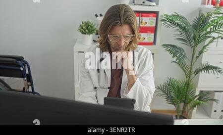 Man doctor in white coat holding tablet with stethoscope, hand on chin pensive pose at desk with computer monitor visible in building; contemplation. Stock Photo