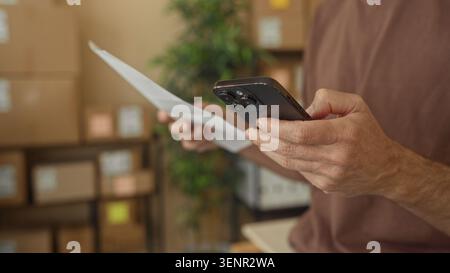 Man holding smartphone and paper in a building, hands visible and stacked boxes nearby; small business focus. Stock Photo