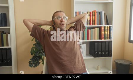 Man with hands behind head leaning back at a desk with book and bookshelf in a building study room wearing glasses and a brown shirt; calm study. Stock Photo