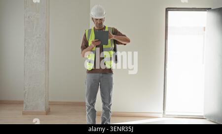 Man in white hardhat and high visibility vest taps tablet and looks up inspecting concrete column in building; focused inspection planning. Stock Photo