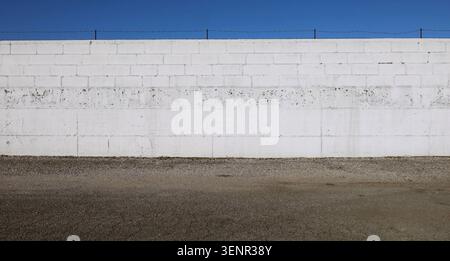 Long white concrete block wall with barbed wire on top. Asphalt street in front, blue sky above. Background for copy space. Stock Photo
