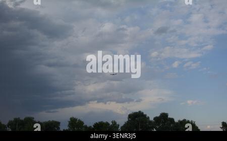 A small airplane flies under a dramatic sky with contrasting gray storm clouds and patches of blue sky visible above the treetops on the horizon. Stock Photo