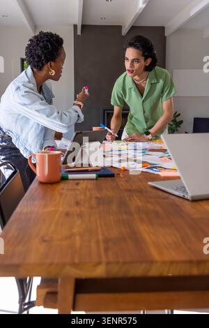 Diverse coworkers collaborating over wood table in office, using pink and blue markers on layouts Stock Photo