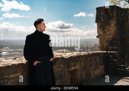 Stylish man in a black coat standing near ancient stone castle ruins with a scenic valley view, bright sky, and cinematic travel atmosphere. Stock Photo