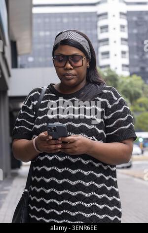 African woman holding smartphone, standing on sidewalk, wearing sunglasses and carrying bag Stock Photo