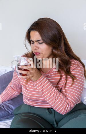African American woman wearing pink-white top and green pants holding amber tea mug on bed Stock Photo