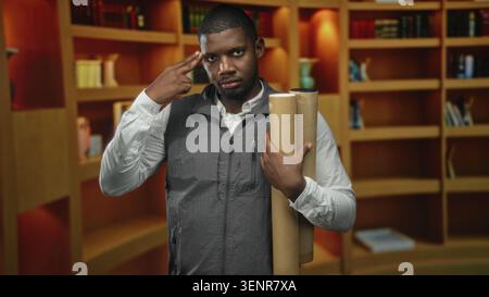 Man holding rolled blueprints and cardboard tubes makes two fingers gesture toward his temple amid wood bookshelves in a building; thoughtful planning Stock Photo