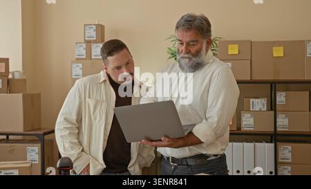 Two men with laptop, one hand on chin and one pointing at screen among stacked parcels in a storage building; teamwork. Stock Photo