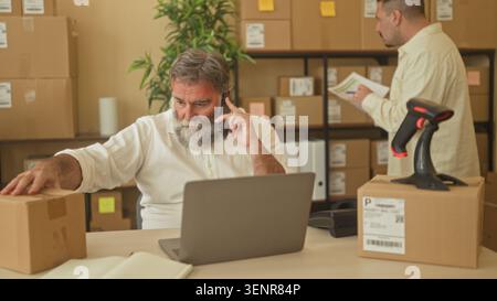 Two men with hand on parcel, holding phone to ear and leaning over laptop in building; teamwork productivity. Stock Photo