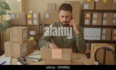 Man talking on smartphone and gesturing with right hand at a packing table in building filled with cardboard boxes wearing green shirt; small business Stock Photo