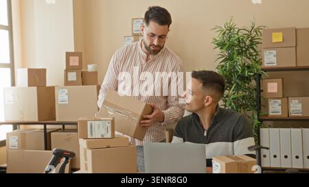 Two men packing boxes, standing man holds a cardboard box while seated man shows phone with hand raised in a building; teamwork productive. Stock Photo