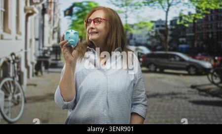 Senior hispanic woman wearing glasses holding a blue piggy bank in her hand on a street with parked cars and bicycle; financial planning hopeful. Stock Photo