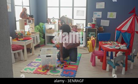 Man kneels assembling and sorting toy blocks into a clear bin on foam number mat inside a kindergarten building; patience nurture. Stock Photo