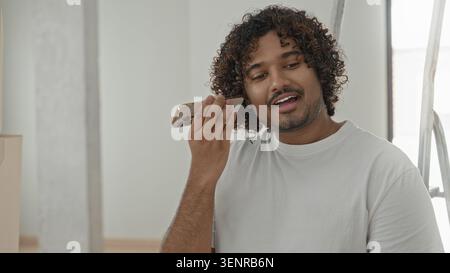 Man holds smartphone to ear and speaks while smiling with curly hair and beard inside a new home building; amused. Stock Photo