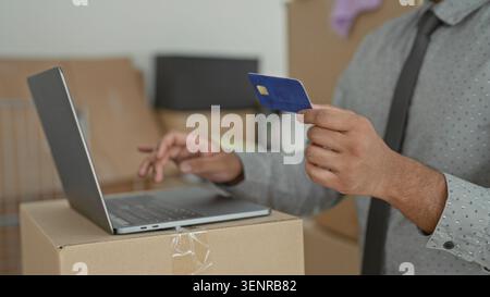 Young indian man holds creditcard in one hand and types on laptop with the other amid moving boxes in a building; convenience. Stock Photo