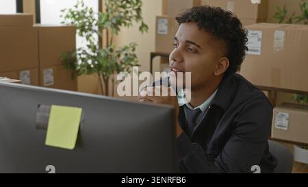 Young man at computer smiles and leans back amid stacked cardboard parcels and shipping boxes on shelving in a building; satisfaction. Stock Photo