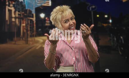 Woman beckons with both hands while smiling on a night street with lampposts and bicycles; welcoming cheerful. Stock Photo