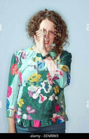 Maggie O'Farrell stands in front of a light blue background. She wears a colorful floral top and holds her hand near her face, smiling slightly at the Stock Photo