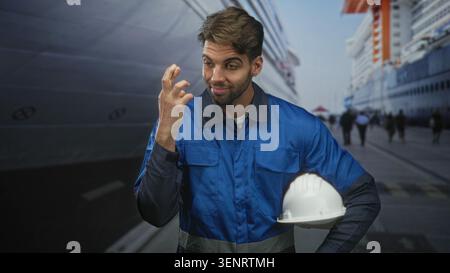 Man construction worker with beard in blue work overalls holding white hardhat at his hip, fingers crossed near face on street beside docked cruise sh Stock Photo