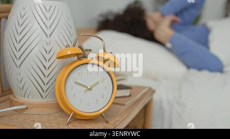 Woman rubbing her eyes beside a yellow alarm clock on a rattan bedside table with books in a building bedroom; sleepy morning. Stock Photo