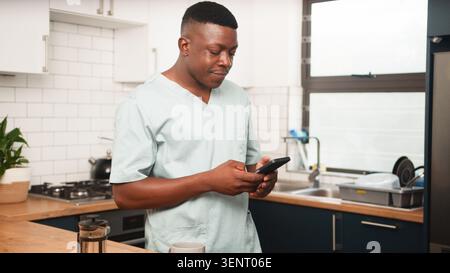 Healthcare Professional In Scrubs Checking Phone In Modern Kitchen During Morning Break Stock Photo