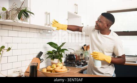 Smiling Young Man Cleaning Kitchen Cabinets in Modern Home Wearing Yellow Gloves and Spray Stock Photo