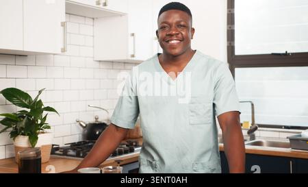 Smiling Male Nurse in Scrubs Standing in Modern Kitchen With Coffee and Plants Stock Photo
