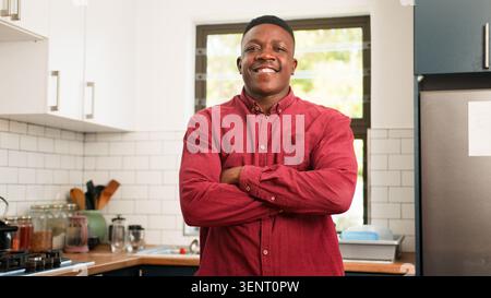 Confident Young Man Smiling With Arms Crossed In Modern Kitchen Wearing Red Shirt Stock Photo