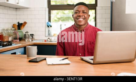 Smiling Young Man Working From Home in Kitchen With Laptop, Coffee and Notepad Stock Photo