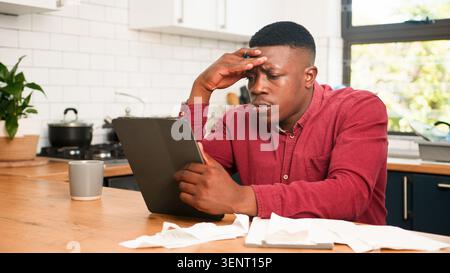 Worried Man Reviewing Bills on Tablet at Home Kitchen Table Managing Personal Finance Stock Photo