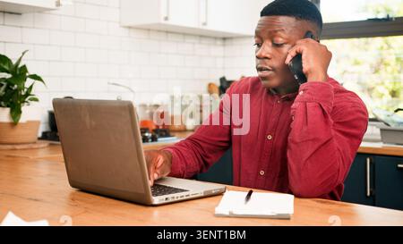 Young Man Working From Home Kitchen Desk On Phone and Laptop Managing Finances Stock Photo